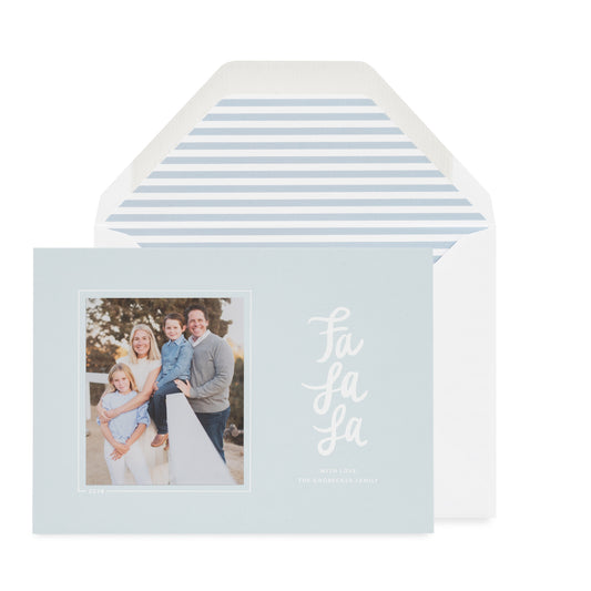 A light blue holiday greeting card with a family photo on the left and red script text reading 'Fa La La' on the right, shown in front of a white envelope with a light blue stripe envelope liner