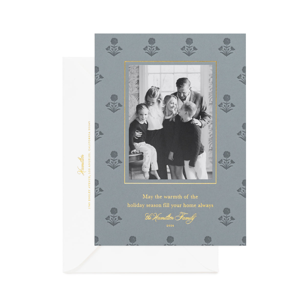 A personalized holiday photo card with a family photo on slate blue paper, featuring gold foil printed with 'may the warmth of the holiday season fill your home always' and dark blue letterpress floral details, with a white envelope