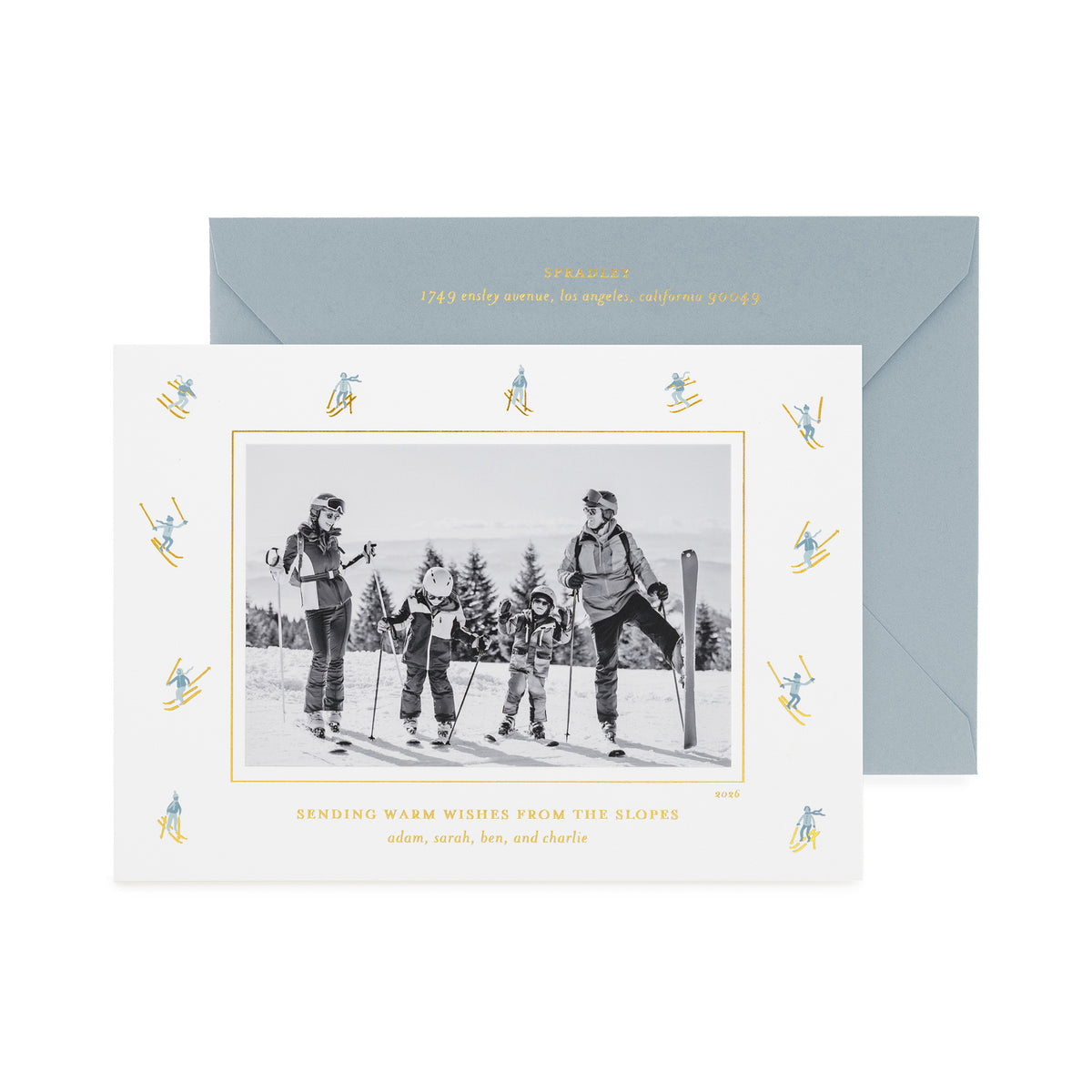 A personalized ski-themed family holiday photo card with a black-and-white family photo in the center, surrounded by a border of small blue and gold skiers, shown with a slate blue envelope with gold foil details and the card printed with 'sending warm wishes from the slopes' 