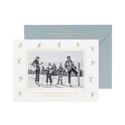 A personalized ski-themed family holiday photo card with a black-and-white family photo in the center, surrounded by a border of small blue and gold skiers, shown with a slate blue envelope with gold foil details and the card printed with 'sending warm wishes from the slopes' 