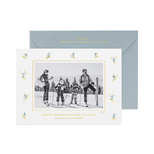 A personalized ski-themed family holiday photo card with a black-and-white family photo in the center, surrounded by a border of small blue and gold skiers, shown with a light blue envelope with gold foil details and the card printed with 'sending warm wishes from the slopes' 
