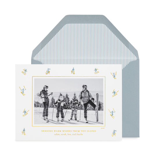 A ski-themed holiday card displayed in front of its matching envelope with blue pinstripe liner. The white card features a central black-and-white photo of a family on the slopes, surrounded by a border of small gold and blue skiers, with the text 'SENDING WARM WISHES FROM THE SLOPES'.