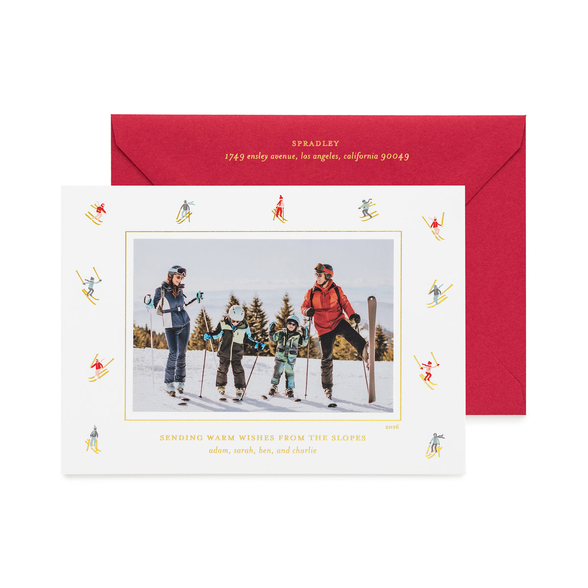A personalized ski-themed holiday photo card in front of its matching red envelope, with a family photo in the center of the card, surrounded by a border of small multicolor and gold skiers, shown with gold foil details and the card printed with 'sending warm wishes from the slopes' 
