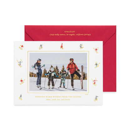 A personalized ski-themed holiday photo card in front of its matching red envelope, with a family photo in the center of the card, surrounded by a border of small multicolor and gold skiers, shown with gold foil details and the card printed with 'sending warm wishes from the slopes' 