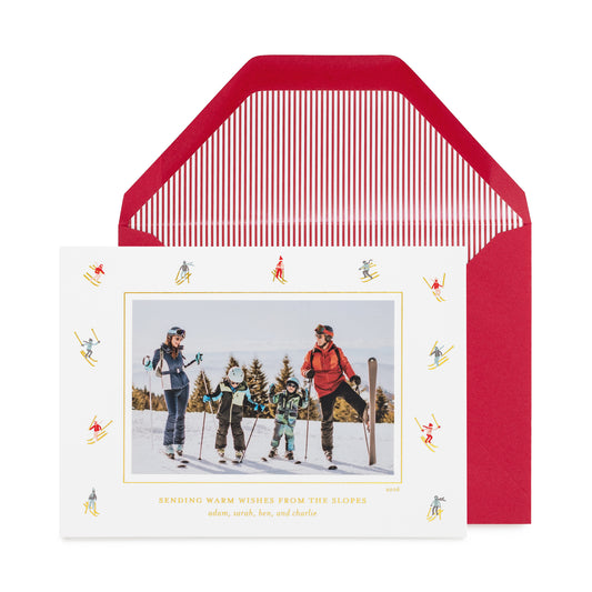 A personalized ski-themed holiday photo card in front of its matching red envelope with a red pinstripe liner, with a family photo in the center of the card, surrounded by a border of small multicolor and gold skiers, shown with gold foil details and the card printed with 'sending warm wishes from the slopes' 