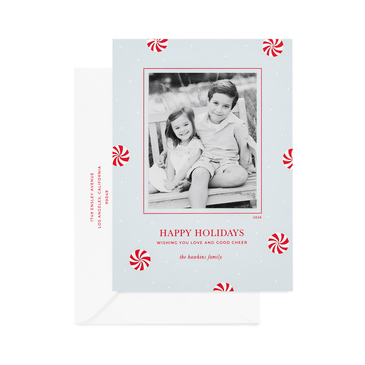 A personalized Christmas photo card on light blue paper with red and white peppermint candy icon pattern, a black-and-white family photo, printed with "merry christmas wishing you love and good cheer" with a white envelope and the address printed in red ink