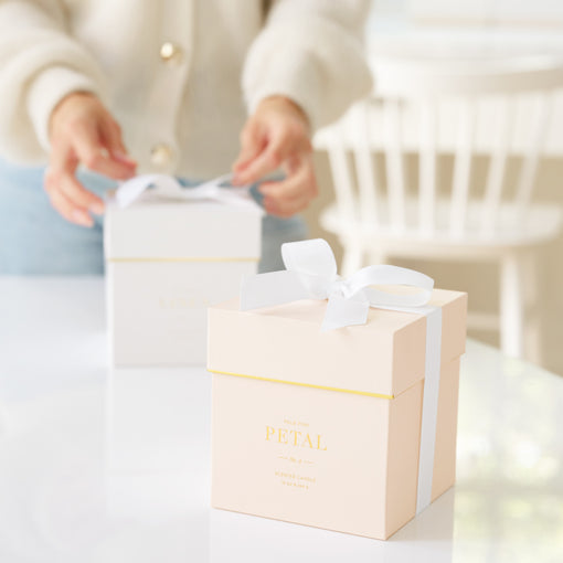 Two candle gift boxes with white ribbons on top on a white table, with a blurred person in the background.