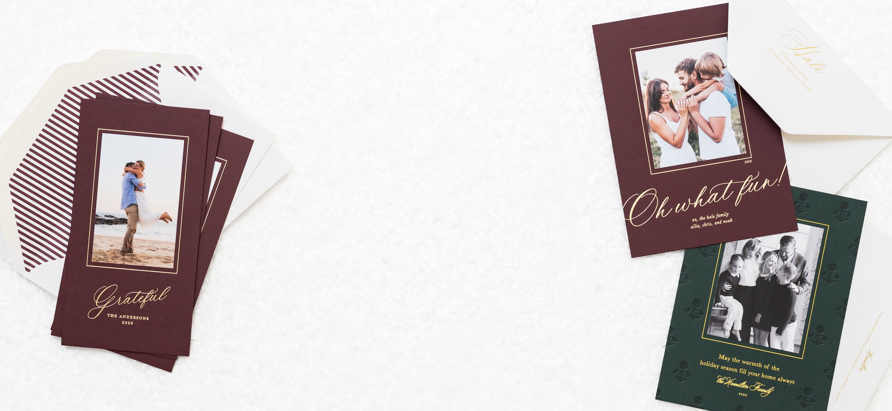 A styled flat-lay of custom photo greeting cards and envelopes on a white textured background. On the left, a stack of maroon cards with gold script lettering and striped envelopes is arranged.