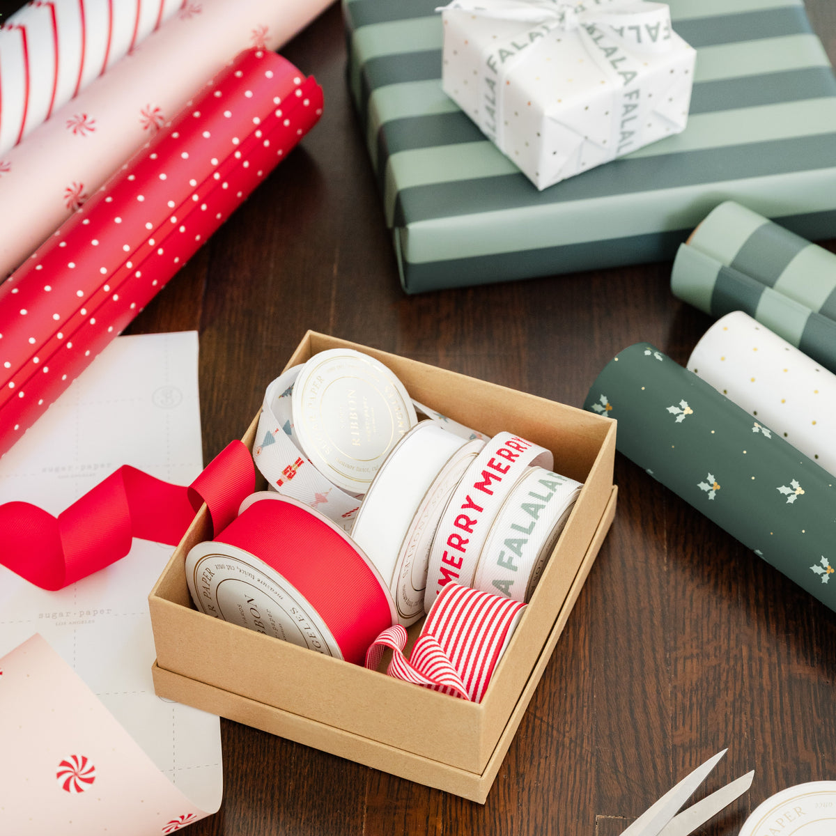 A styled flat-lay of holiday gift wrapping supplies on a dark wood table, featuring a box of different ribbon rolls with red and green patterns, rolls of coordinating wrapping paper, gold scissors, and a stack of green wrapped gift boxes