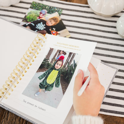 A hand holds a white pen over an open spiral photo album with gold rings, showing a printed page titled 'Halloween Memories' with a child in a green dinosaur costume. The scene is styled on a striped fabric surface with other printed photos and white decorative pumpkins