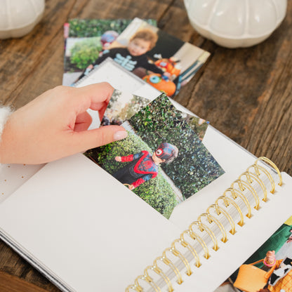 A hand placing printed Halloween costume photos into a white spiral photo album with gold rings on a rustic wooden table, with other printed photos and pumpkins in background