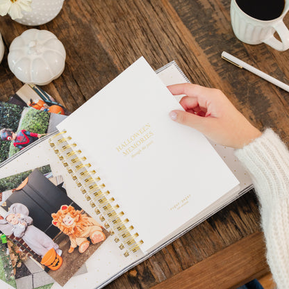 A hand wearing a white knit sweater holds an open white spiral-bound journal on a rustic wooden table. The cover reads 'HALLOWEEN MEMORIES' in gold lettering with photos of kids in halloween costumes and pumpkins in the background
