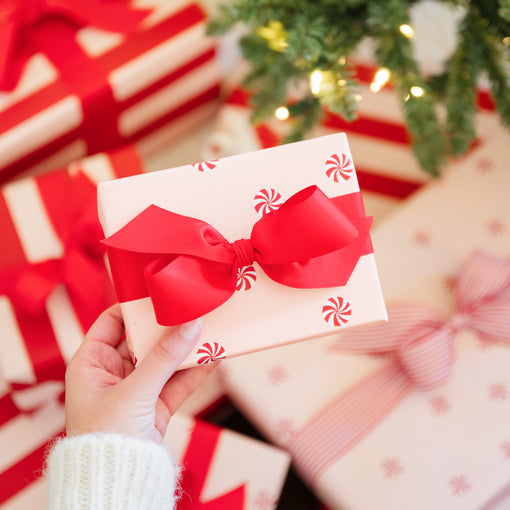A hand in a white knit sweater holds a small gift box wrapped paper with red candy cane prints and a large red ribbon bow. Other wrapped presents and a Christmas tree with warm lights are blurred in the background.