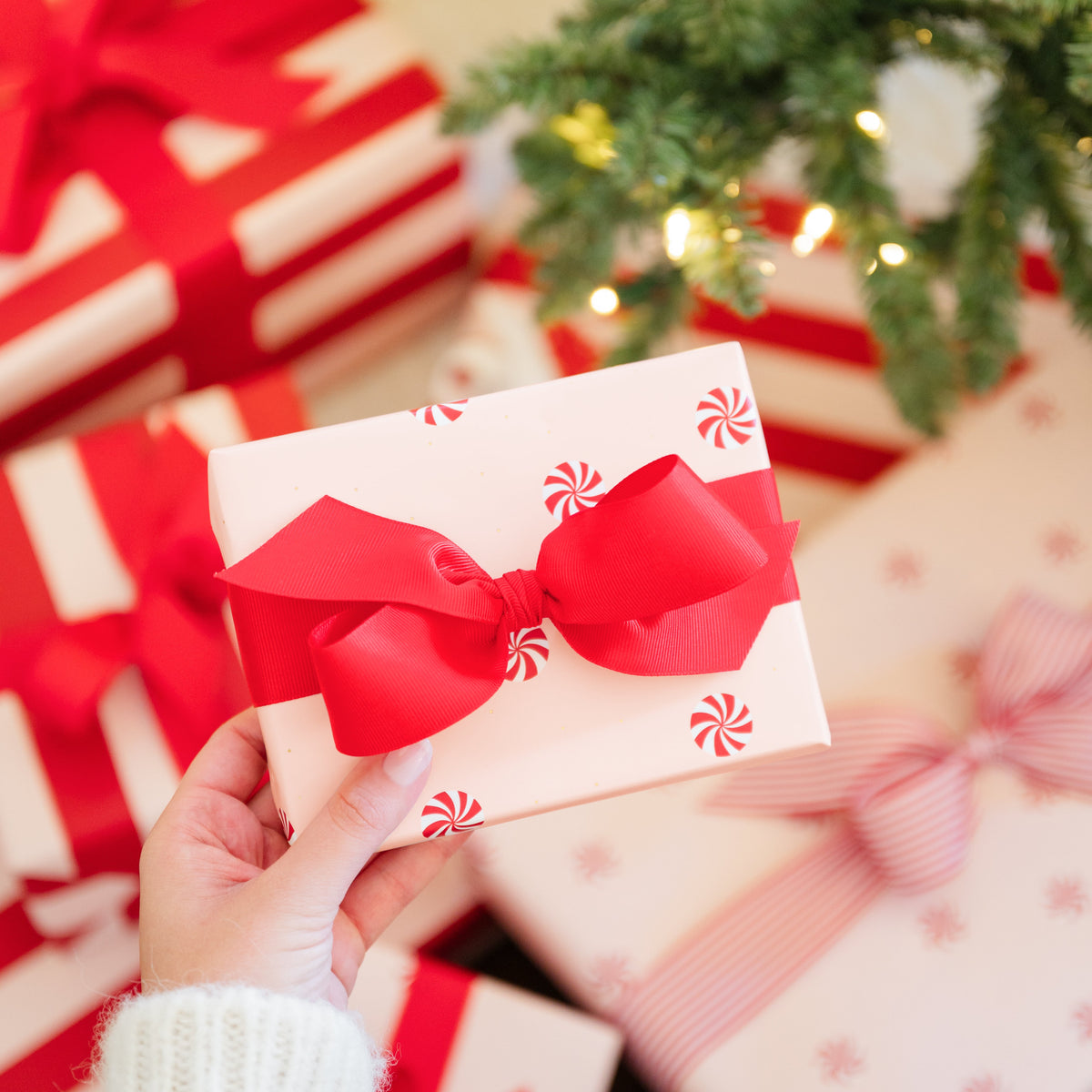 A hand in a white knit sweater holds a small wrapped gift box with a large red bow and red peppermint candy print. Other wrapped Christmas presents with red and white stripes and bows are scattered around under the green branches of a lit Christmas tree.