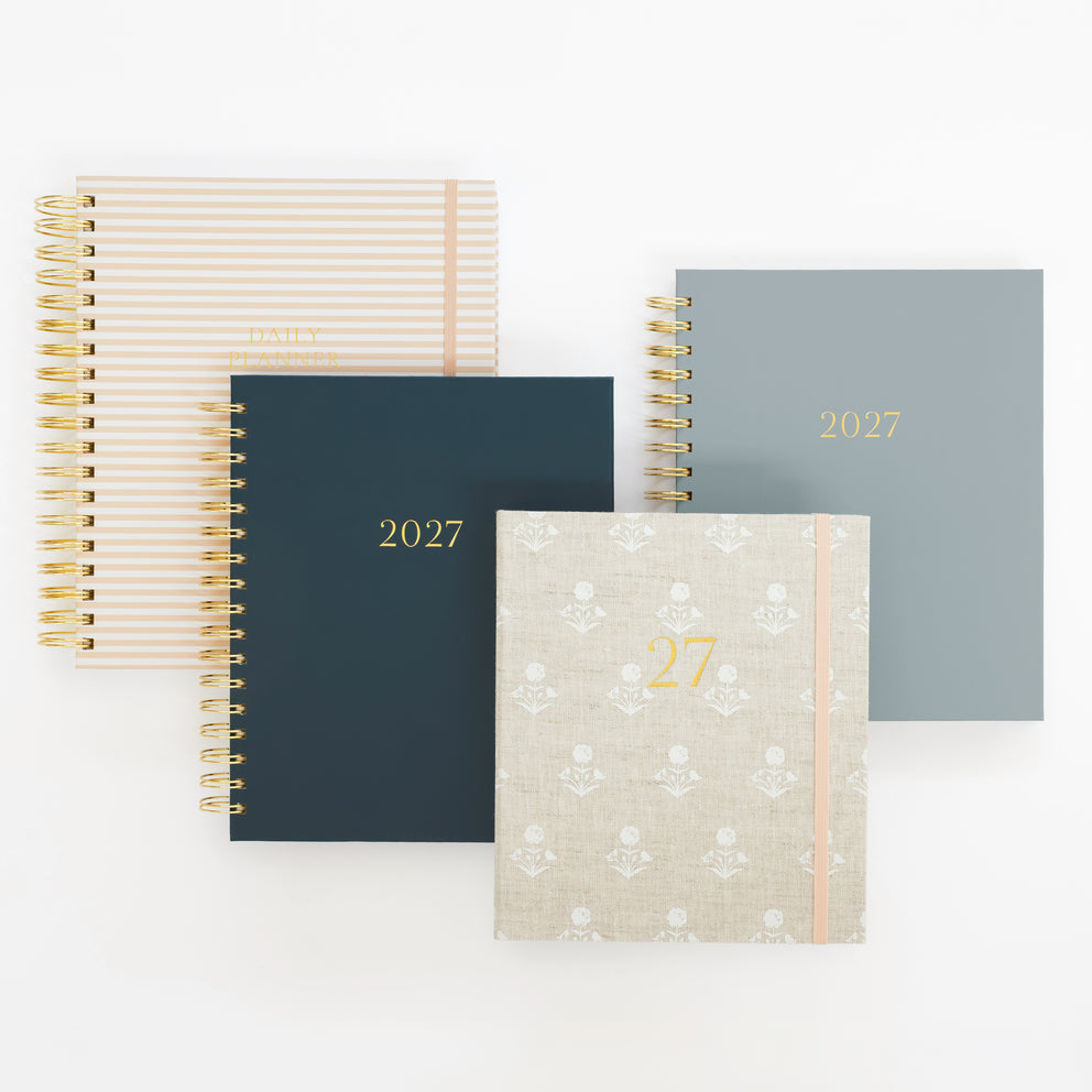 Four 2027 planners arranged on a clean white background. The set includes four designs: a white and pale pink striped cover, a dark navy blue cover with gold text, a dusty blue cover with gold text, and a light beige cover with a subtle floral pattern and gold text and a pink elastic ribbon