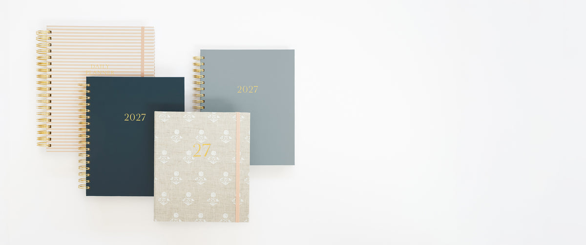 Four 2027 planners arranged on a clean white background. The set includes four designs: a white and pale pink striped cover, a dark navy blue cover with gold text, a dusty blue cover with gold text, and a light beige cover with a subtle floral pattern and gold text and a pink elastic ribbon