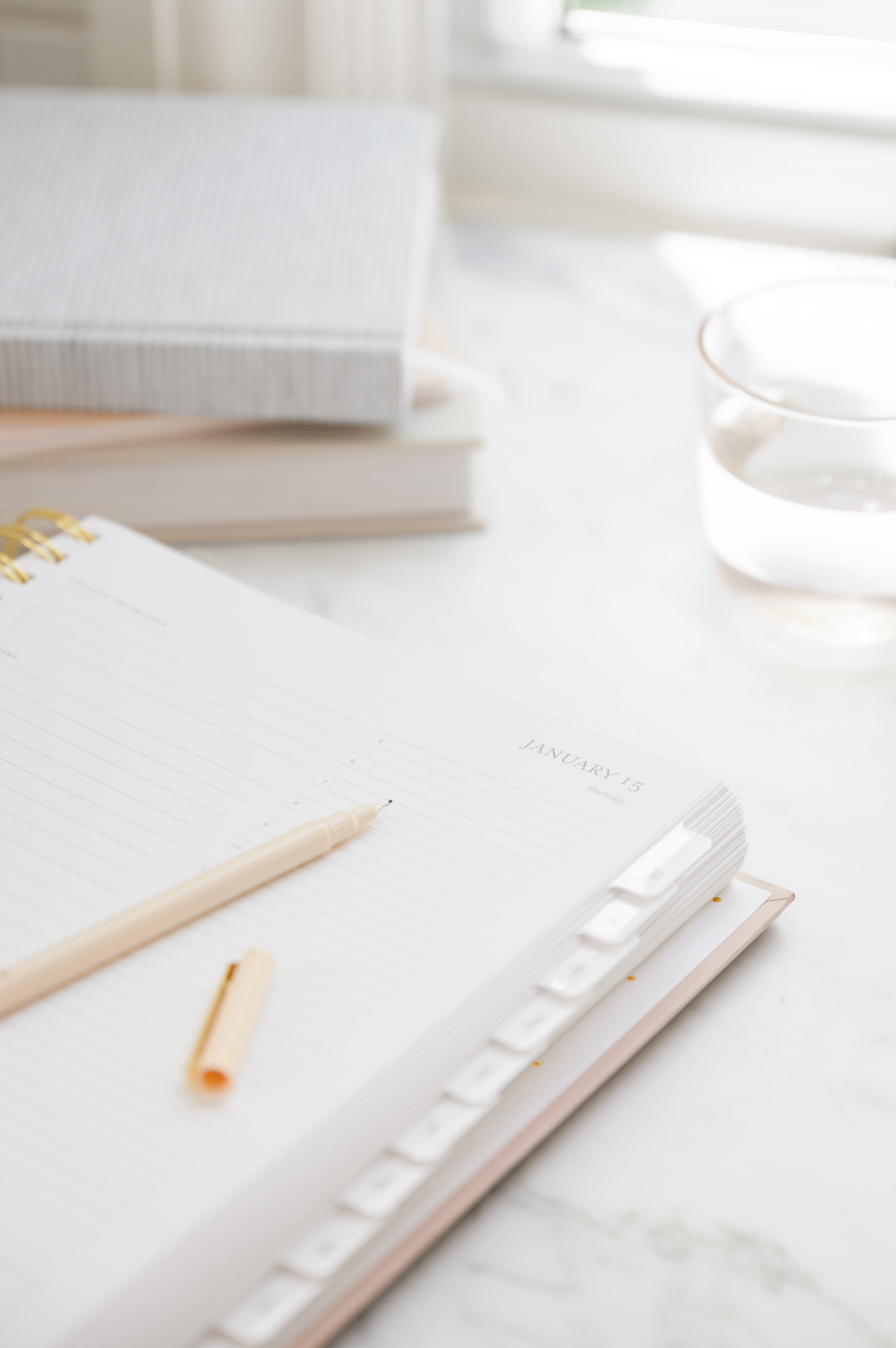 An open lined planner with a gold pen and pen cap on a light marble surface, with stacked journals and a glass of water softly blurred in the background.