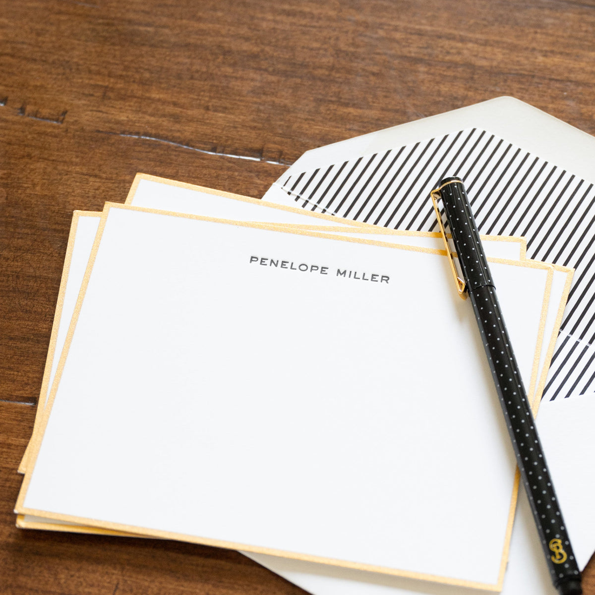 A styled flat-lay of stationery on a rustic wooden table, featuring a stack of white cards with gold borders printed with black ink, a white envelope with black diagonal stripes, a black pen with gold lettering