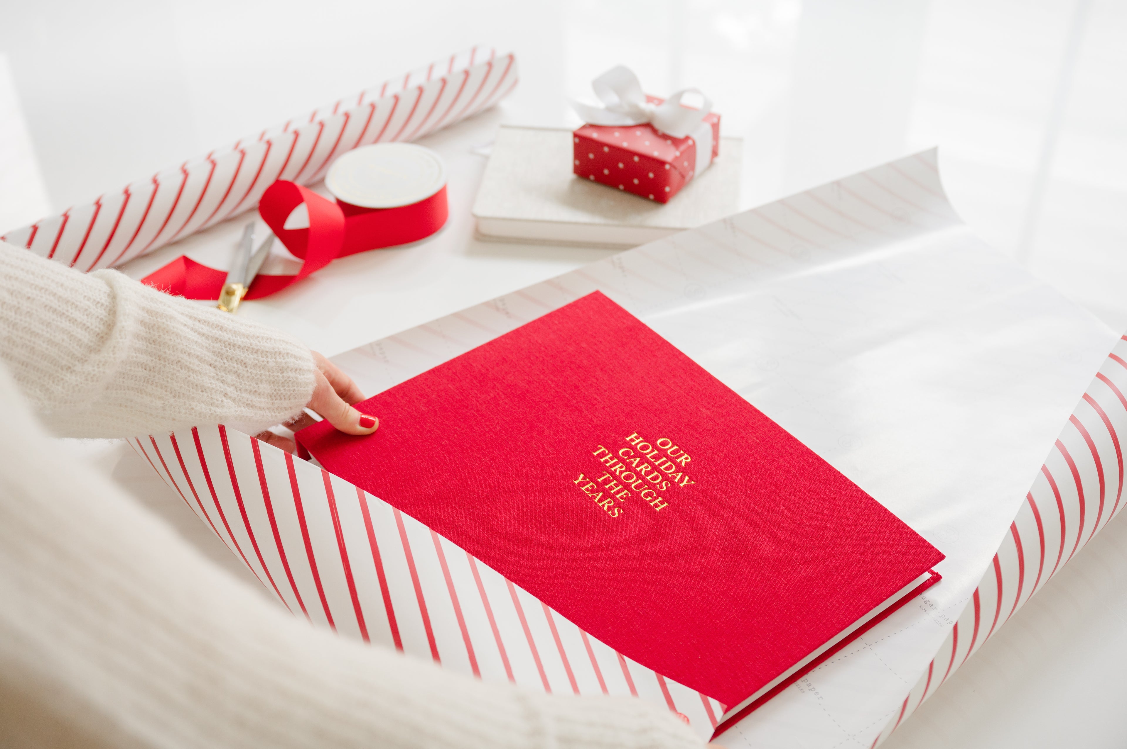 Person wrapping a red holiday card book with 'our holiday cards through the years' with a red stripe wrapping paper roll, red ribbon, scissors and a small red with white dot wrapped box