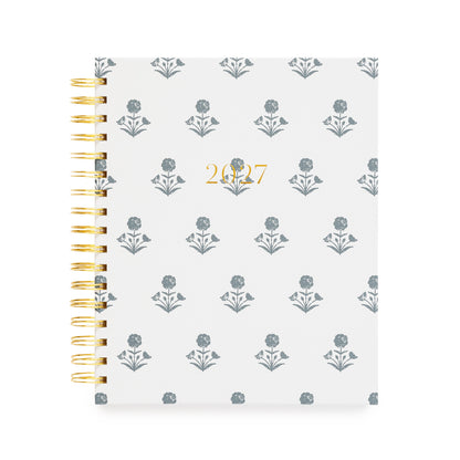 A 2027 spiral-bound planner with a white cover with a blue floral pattern and gold foil text reading '2027' in the center. The left side features a gold-tone spiral coil binding.