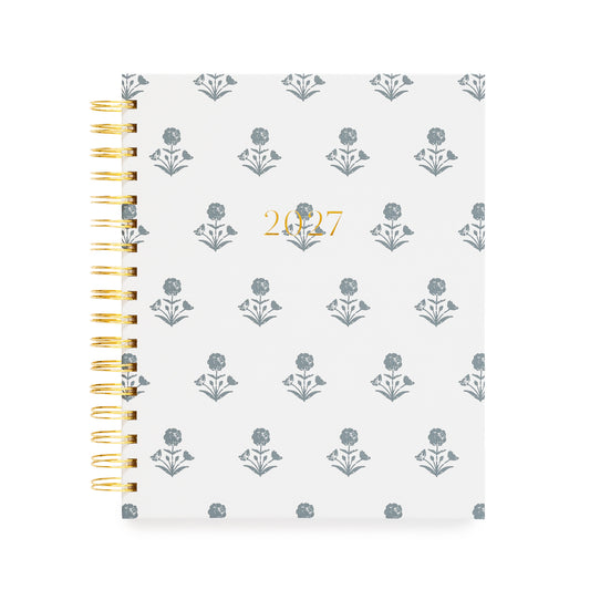 A 2027 spiral-bound planner with a white cover with a blue floral pattern and gold foil text reading '2027' in the center. The left side features a gold-tone spiral coil binding.