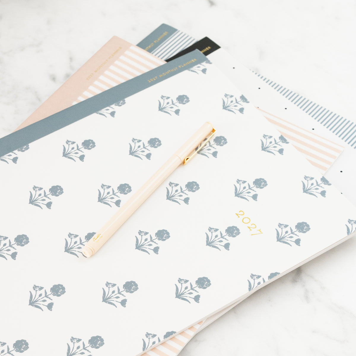 A 2027 planner with a white cover decorated with a blue-gray floral pattern and gold '2027' text, styled on a marble surface with a gold pen and other flat monthly planners underneath.