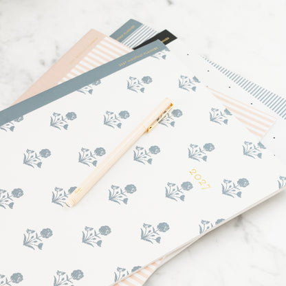 A 2027 planner with a white cover decorated with a blue-gray floral pattern and gold '2027' text, styled on a marble surface with a gold pen and other flat monthly planners underneath.