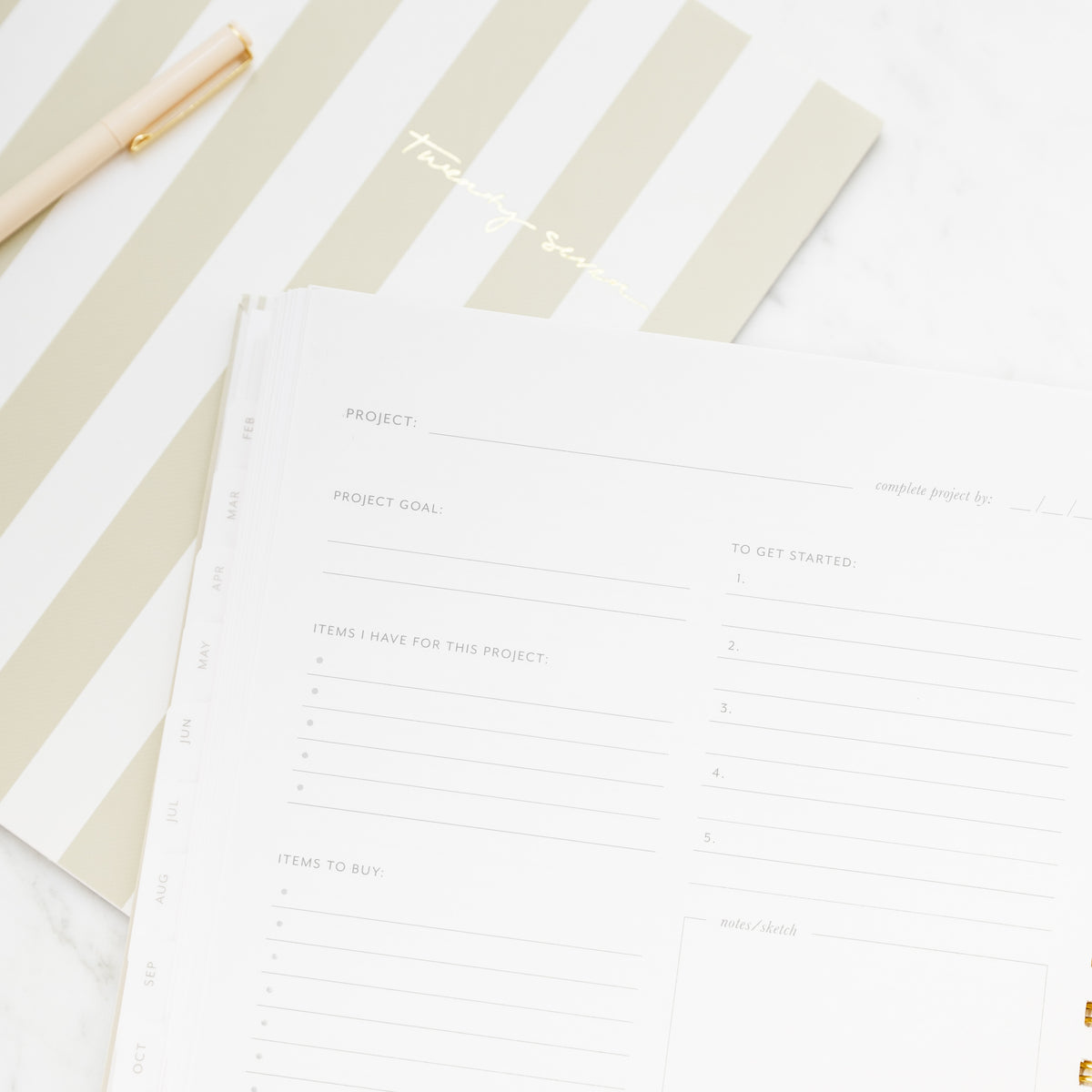 A close-up of a spiral-bound planner with gold wire rings, showing printed project planning pages with goals, items to have, items to buy, to get started and notes