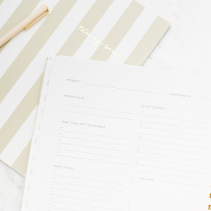 A close-up of a spiral-bound planner with gold wire rings, showing printed project planning pages with goals, items to have, items to buy, to get started and notes