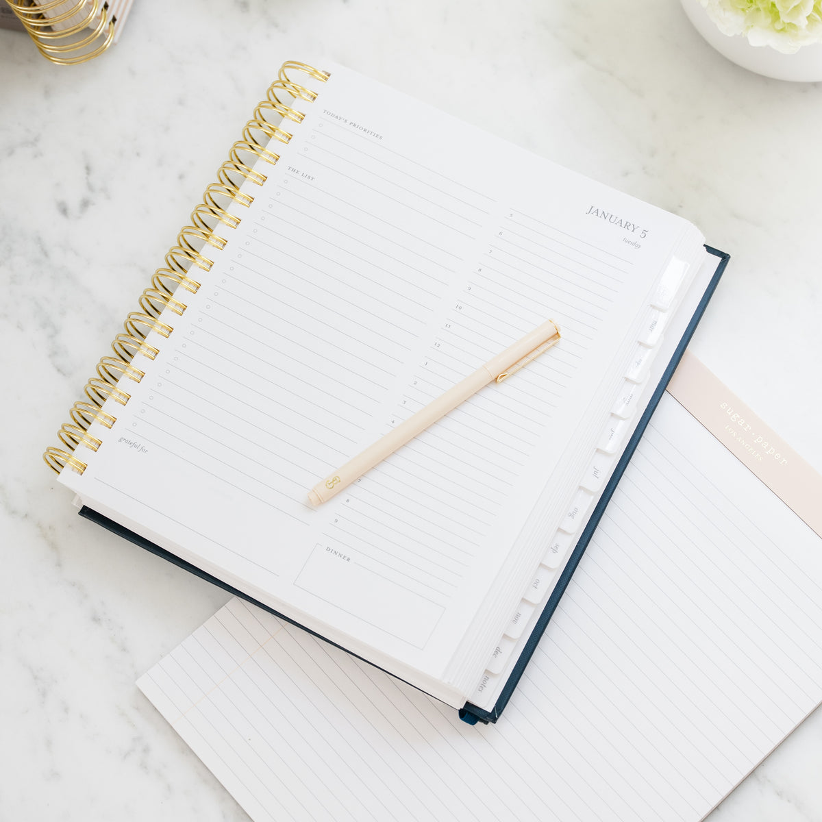 An open spiral-bound daily planner with a gold wire binding showing january daily planning page with times of day, priorities, the list, grateful for, etc