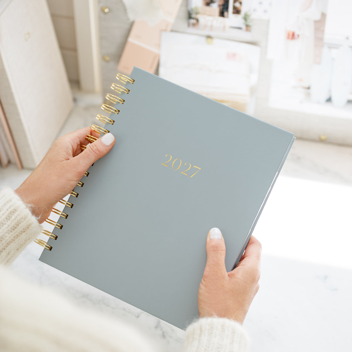 A person holding a 2027 spiral-bound planner with gold lettering and gold wire binding, set against a bright, softly blurred indoor background.