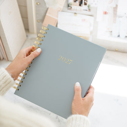 A person holding a 2027 spiral-bound planner with gold lettering and gold wire binding, set against a bright, softly blurred indoor background.
