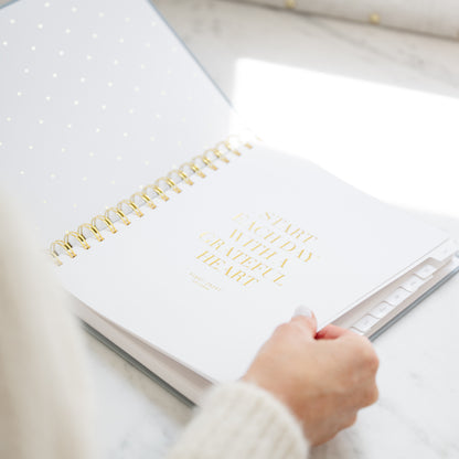 A white spiral-bound planner with gold foil text reading 'START EACH DAY WITH A GRATEFUL HEART' on the cover, shown open on a light marble surface with a hand turning the page.
