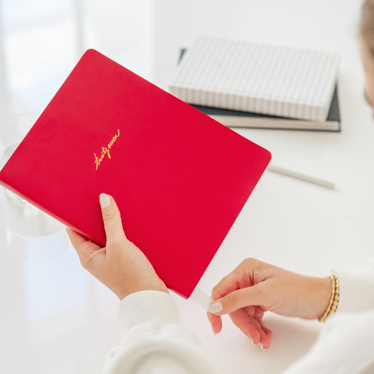 A person holding a bright red notebook with gold script lettering on the cover, photographed in a bright, clean indoor setting with a white desk and blurred background items.