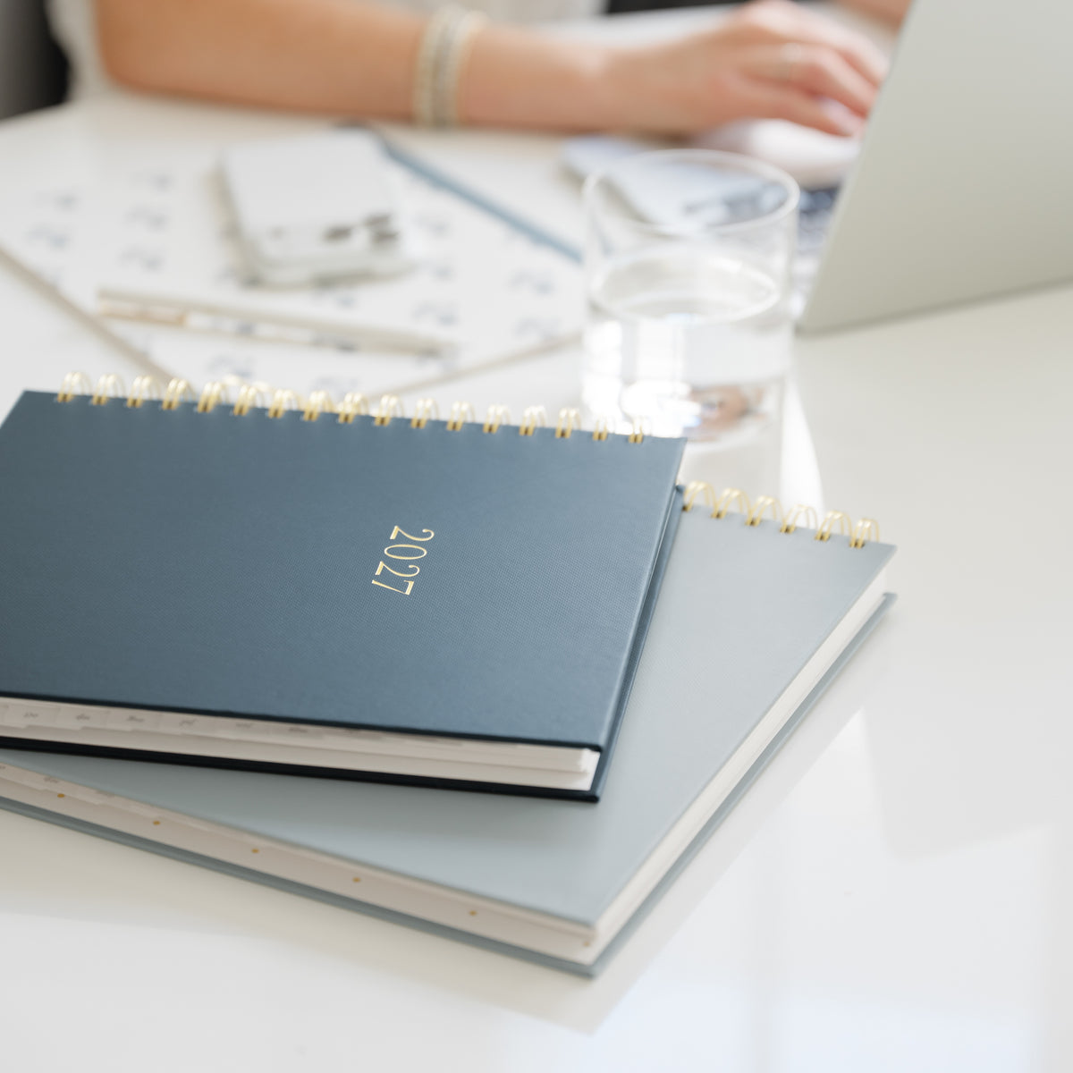 A navy blue 2027 spiral-bound planner resting on top of a gray notebook on a white desk, with a blurred office scene in the background including a person's hands and a laptop.