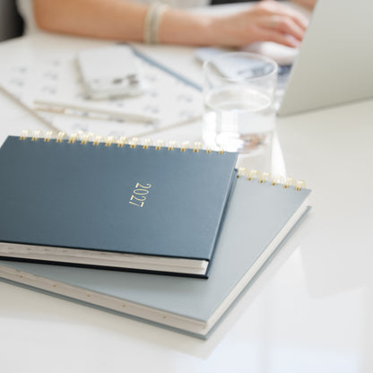 A navy blue 2027 spiral-bound planner resting on top of a gray notebook on a white desk, with a blurred office scene in the background including a person's hands and a laptop.