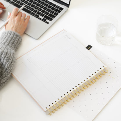 A top-down view of a person's hand resting on a laptop next to an open spiral-bound planner with gold rings and a white page, on a white desk with a glass of water.