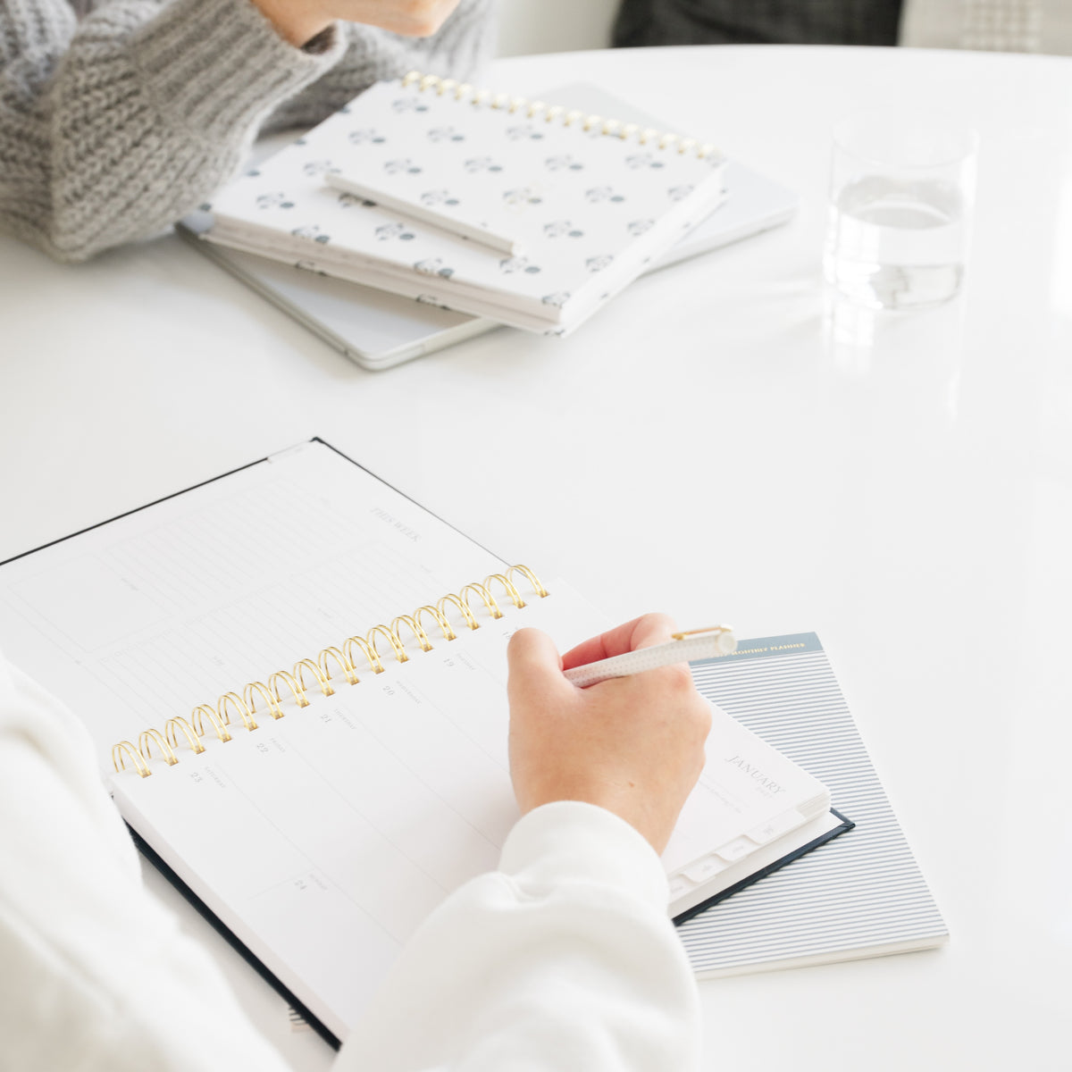 A person writing in a white spiral-bound planner with a gold coil on a white table, with other planners and a glass of water in the background.