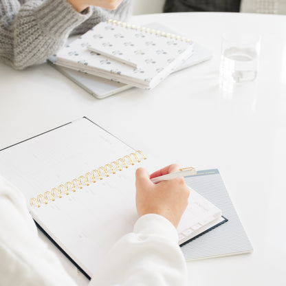 A person writing in a white spiral-bound planner with a gold coil on a white table, with other planners and a glass of water in the background.