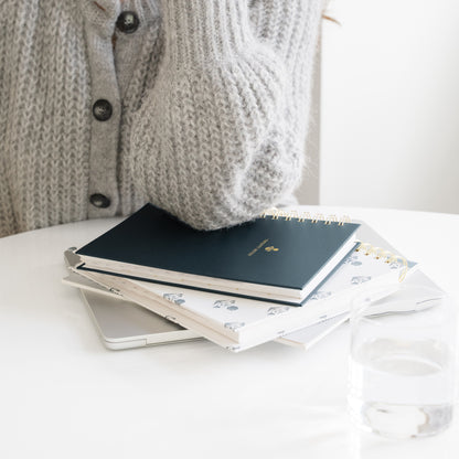 A stack of planners and a silver laptop on a white table, with a person wearing a gray knit cardigan resting their arm on top of the planners