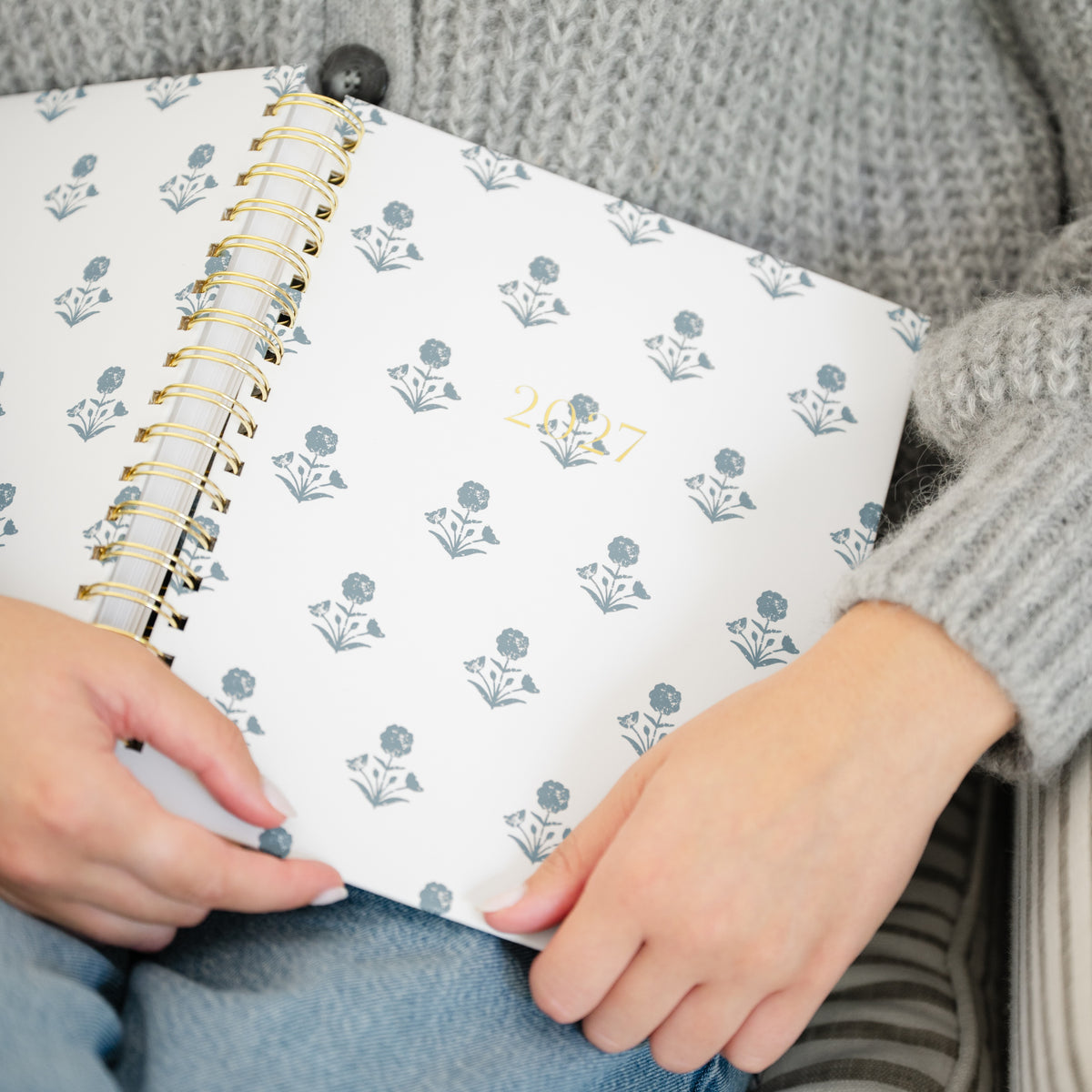A close-up of a person holding an open spiral-bound planner. The cover is white with a repeating muted blue floral pattern and gold-toned spiral binding.
