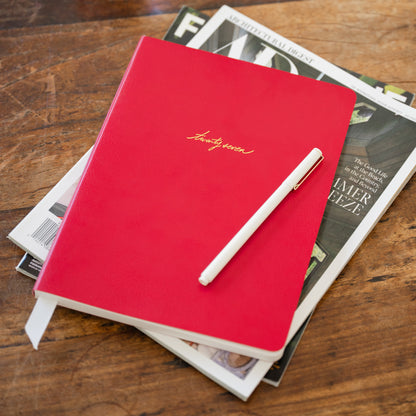 A red soft leather agenda with gold foil script reading 'twenty seven' sits on top of stacked magazines on a wooden table, with a white pen resting on top