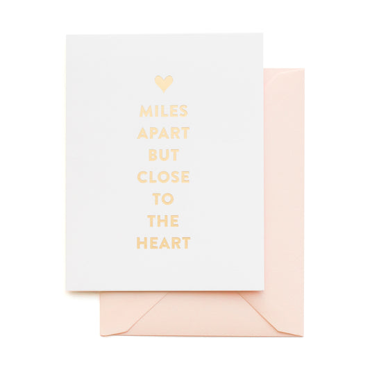 Greeting card with 'Miles apart but close to the heart' text with gold foil on a white background with a pink envelope.