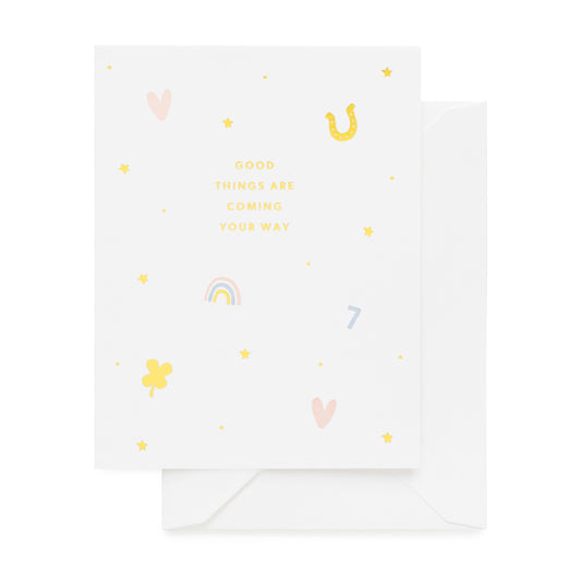 White greeting card with gold foil, pink, and blue icons in heart, horseshoe, rainbow, and lucky charm printed with 'good things are coming your way'