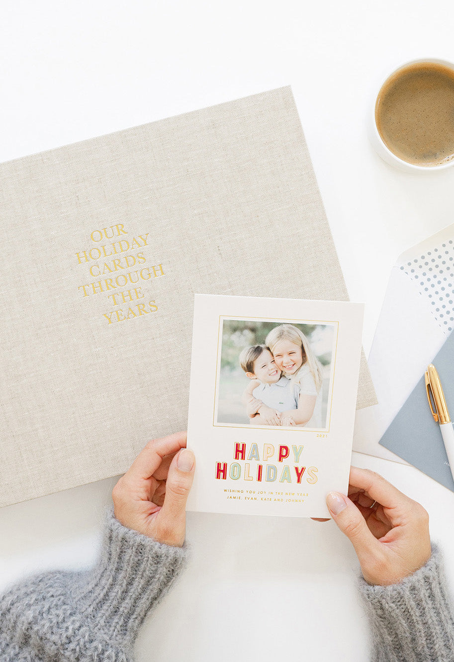 Person holding a photo card with 'Happy Holidays' text, with a holiday card keepsake book and coffee cup on a light surface.