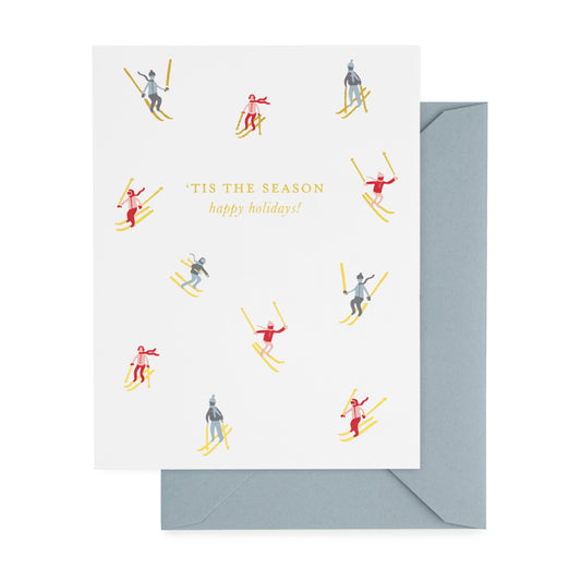 A white greeting card with the text 'TIS THE SEASON happy holidays!' and small illustrated skiers in red, blue, and gold foil, shown with a light blue envelope