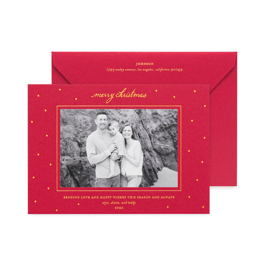 A red personalized Christmas photo card with gold text and gold dot accents, shown in front of a matching red envelope. The card features a black-and-white photo of a family of three, with gold script reading 'Merry Christmas' at the top and 'sending love and happy wishes this season and always'