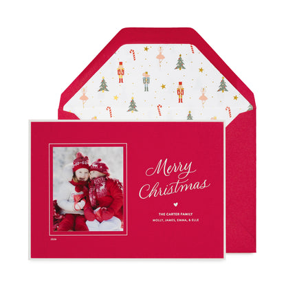 A personalized Christmas photo card on a red card featuring a photo of a family in winter clothing, the text 'Merry Christmas' in white script, and the names 'THE CARTER FAMILY' with a small heart and a red envelope with a nutcracker icon pattern liner.