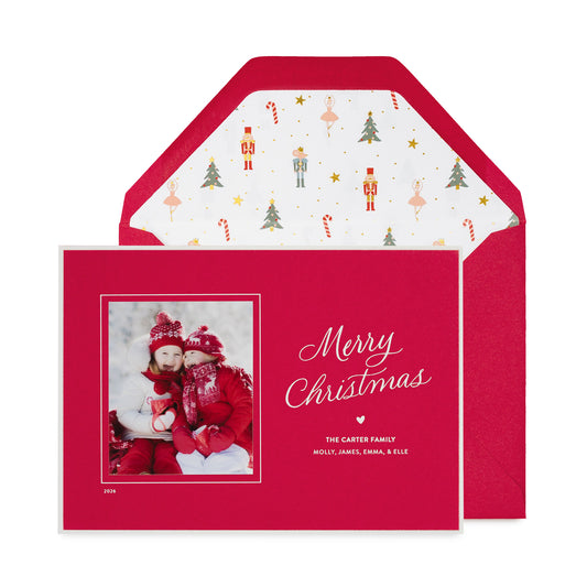A personalized Christmas photo card on a red card featuring a photo of a family in winter clothing, the text 'Merry Christmas' in white script, and the names 'THE CARTER FAMILY' with a small heart and a red envelope with a nutcracker icon pattern liner.