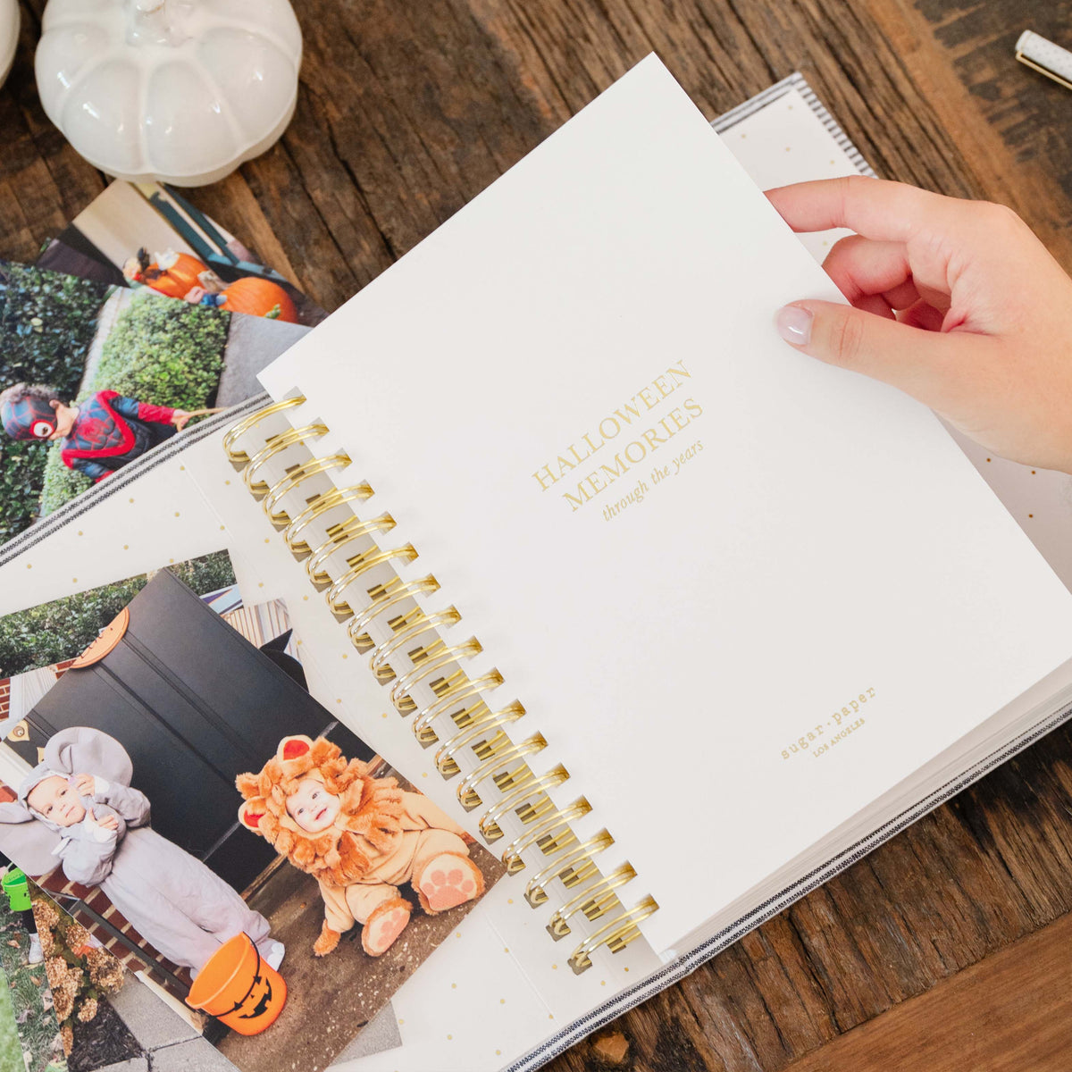 A hand holding an open white spiral-bound journal titled 'HALLOWEEN MEMORIES through the years' on a rustic wooden table, with photos and a white pumpkin visible in the background.